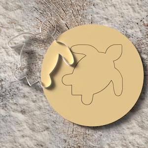 Puede incluir: Un cortador de galletas circular de color beige claro con un contorno de tortuga, junto a un cortador de galletas transparente en forma de tortuga. Los artículos están sobre una superficie de vetas de madera. El diseño de la tortuga es un tema común para hornear.