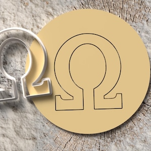 May include: A clear plastic Omega symbol cookie cutter next to a tan circle with a black Omega symbol outline. The objects are on a textured, light brown wooden surface.