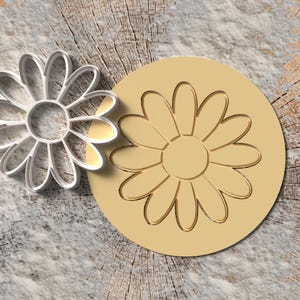 Peut inclure: Un emporte-pièce en forme de marguerite argenté à côté d'un tampon circulaire beige avec un motif de marguerite. Les articles sont sur une surface texturée et claire avec un fond en grain de bois. L'emporte-pièce est en métal.