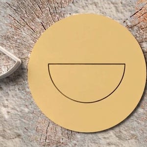 Peut inclure: Un emporte-pièce en forme de demi-cercle en plastique blanc, placé à côté d'une découpe de pâte circulaire jaune clair qui présente un contour en demi-cercle imprimé au centre, créant une empreinte nette de la forme sur la surface plane.