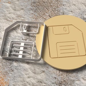 May include: A beige floppy disk cookie cutter and a matching cookie. The cookie cutter is a white plastic outline of the floppy disk shape. The cookie is a solid beige color with the floppy disk design embossed on it. The items are on a wood surface.