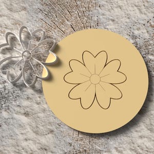 Puede incluir: Un cortador de galletas plateado con forma de flor junto a un círculo de color beige con un contorno de flor. Los objetos están sobre una superficie con vetas de madera.