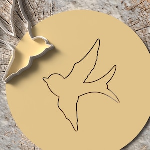 Könnte beinhalten: Ein goldfarbenes, vogelförmiges Design mit weißem Umriss befindet sich in der Nähe eines beigefarbenen Kreises mit einem Vogelumriss. Der Hintergrund ist eine strukturierte, hellbraune Holzoberfläche. Das Vogeldesign ist eine stilisierte Darstellung einer Schwalbe.