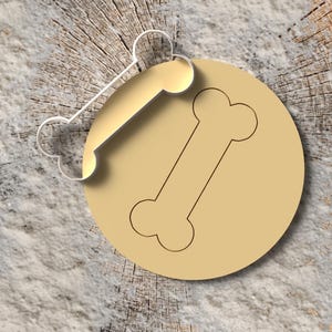 Könnte beinhalten: Ein Hundeknochen-Ausstecher und ein Knochen-Design auf einem beigefarbenen Kreis. Der Ausstecher ist aus hellem Metall gefertigt. Der Hintergrund ist eine Holzscheibe.