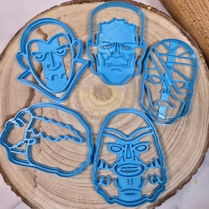Könnte beinhalten: Fünf blaue Ausstechformen aus Kunststoff in Form klassischer Filmmonster, darunter ein Vampir, Frankensteins Monster, die Mumie, die Kreatur aus der schwarzen Lagune und ein Werwolf. Die Ausstecher liegen auf einer Holzoberfläche.