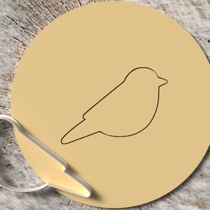 May include: A bird-shaped cookie cutter next to a tan circle with a bird outline. The cookie cutter is silver-colored. The background is a wood-grain pattern.