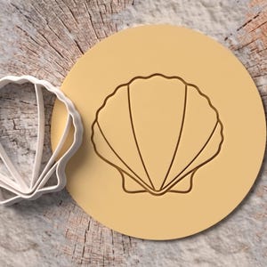 Op de afbeelding: Een witte plastic koekjesvorm in de vorm van een schelp naast een rond stuk lichtbruin deeg met een afgedrukte schelpomtrek. De vorm heeft een geschulpte rand en interne ribbels die een gedetailleerde afdruk van een waaiervormige schelp maken.