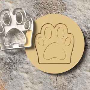 May include: A clear plastic paw print cookie cutter next to a tan-colored paw print stamp. The paw print design is a popular theme for pet-related products. The items are set against a textured, wood-grain background.