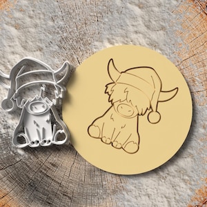 Puede incluir: Un cortador de galletas de metal y un sello con una vaca de las Tierras Altas con un gorro de Papá Noel. El cortador de galletas es plateado, y el sello es un círculo marrón claro con un contorno detallado de la vaca. Los artículos están sobre una superficie de madera.