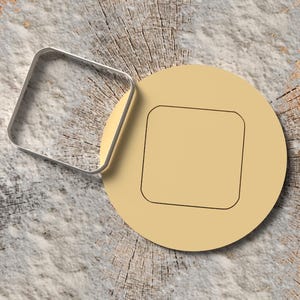 May include: A silver square cookie cutter next to a tan circular cookie with a square indentation. The objects are on a light grey and brown wood slice surface. The cookie cutter is made of shiny metal.