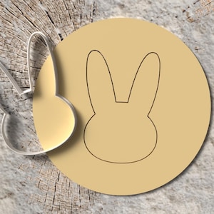 Könnte beinhalten: Ein klarer, hasenförmiger Ausstecher aus Kunststoff neben einem hellbraunen Kreis mit einer Hasenkontur. Die Gegenstände befinden sich auf einer strukturierten, hellbraunen und grauen Oberfläche, was auf ein rustikales oder natürliches Thema hindeutet. Das Design ist einfach und sauber.