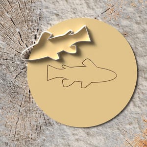 Könnte beinhalten: Goldfarbener Fisch-Ausstecher mit passender Kontur auf einem beigefarbenen Kreis. Der Hintergrund ist eine verwitterte Holzoberfläche, ideal für Back- oder Bastelprojekte mit Fischerei- oder Naturmotiven.