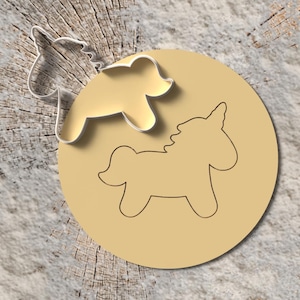 May include: A unicorn-shaped cookie cutter and a unicorn-shaped imprint on a tan circle. The cookie cutter is made of silver metal. The unicorn imprint is on a tan circle. The background is a light brown wood.
