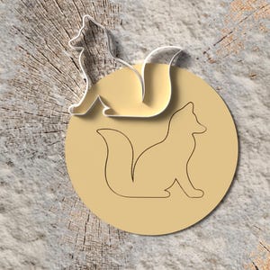 Könnte beinhalten: Ein hellbrauner fuchsformiger Anhänger mit einem Ausschnittdesign, der auf einer hellbraunen kreisförmigen Basis sitzt. Ein passender fuchsformiger Ausschnitt befindet sich über der Basis. Der Hintergrund ist eine strukturierte, hellbraune Oberfläche.