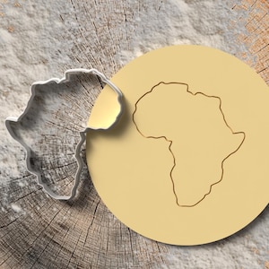 May include: A silver cookie cutter shaped like Africa, placed beside a round, pale yellow biscuit with the continent's outline. The items rest on a textured, light brown wooden surface.