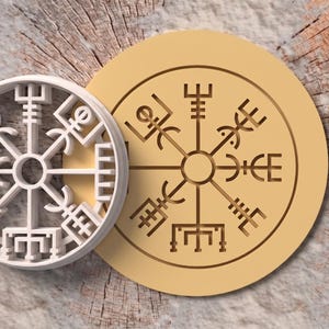 Op de afbeelding: Een witte ronde koekjesvorm met het Vegvisir-symbool met acht uitstralende staven en een bijpassend lichtbruin rond koekje met hetzelfde ingewikkelde runenkompasontwerp, voorzien van duidelijke geometrische lijnen en patronen.