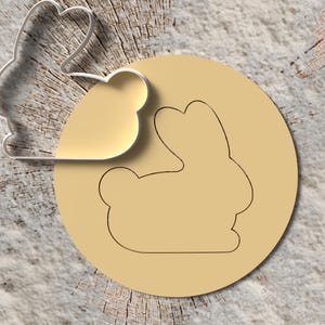 Könnte beinhalten: Ein silberfarbener, hasenförmiger Ausstecher neben einem beigefarbenen Kreis mit einer Hasenkontur. Das Set befindet sich auf einer strukturierten, hellbraunen Oberfläche, passend zum Backen.