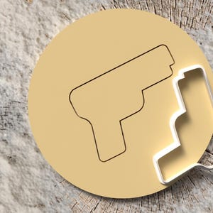 Puede incluir: Un cortador de galletas circular de color beige con un contorno de pistola y un cortador de metal con forma de pistola. La imagen está sobre una superficie de madera clara y texturizada.