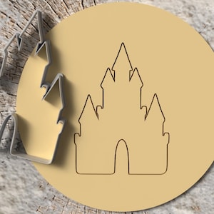 May include: A castle-shaped cookie cutter next to a cookie with a castle outline. The cookie cutter is silver, and the cookie is light brown. The background is a wooden surface.
