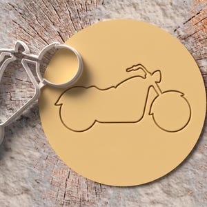 May include: A white plastic motorcycle cookie cutter positioned next to a circular piece of light tan dough that has been embossed with the same motorcycle outline. The design features a classic cruiser motorcycle silhouette with handlebars.