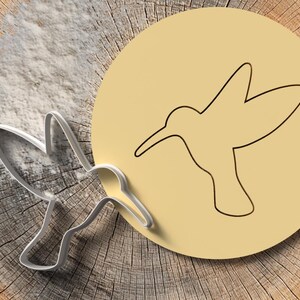 Może przedstawiać: Metalowa foremka do ciastek w kształcie kolibra obok żółtego koła z konturem kolibra. Obiekty znajdują się na rustykalnej, drewnianej powierzchni z warstwą białego proszku, co sugeruje pieczenie.