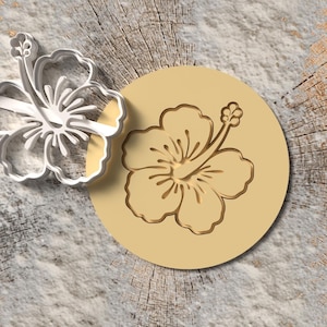 Puede incluir: Un cortador de galletas plateado con forma de flor de hibisco junto a un sello circular de color beige con un diseño de flor de hibisco a juego. Los objetos están sobre una superficie de vetas de madera.