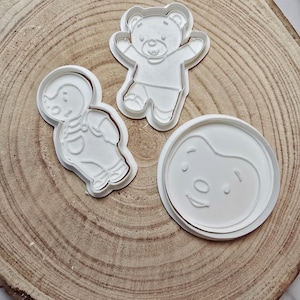 Peut inclure: Trois emportes-pièces en plastique blanc en forme d'ours, de personne et de visage rond. L'ours est debout, les bras tendus. La personne porte une salopette. Le visage rond a un simple sourire et un nez.