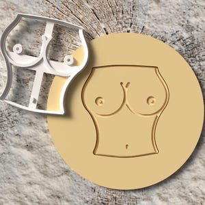 Könnte beinhalten: Ein Ausstecher und ein Keksstempel, beide in Form eines Torsos. Der Ausstecher ist weiß und der Stempel ist beige. Der Keksstempel befindet sich auf einem beigen Kreis. Der Hintergrund ist eine Holzmaserung.