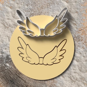 Op de afbeelding: Een koekjessnijdersset met engelen vleugels. De set bevat een metalen omtrek van de vleugels en een bijpassende afdruk op een gele cirkel. De items worden gepresenteerd op een houten schijf achtergrond.