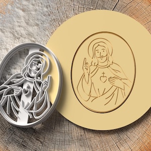 May include: A silver-colored cookie cutter and a tan-colored stamp, both featuring an oval design of a religious figure with a halo, a heart, and a cross. The items are displayed on a wooden surface.