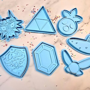 Puede incluir: Un conjunto de seis cortadores de galletas de plástico azul con forma de escudo, diamante, cabeza de lobo, triángulo, instrumento musical y criatura alada.