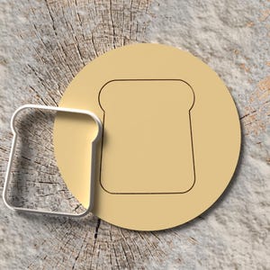 Puede incluir: Un cortador de galletas con forma de pan y una plantilla a juego se muestran sobre una superficie de madera. El cortador de galletas es de metal plateado y la plantilla es de color beige claro. El diseño es un simple contorno de una rebanada de pan.