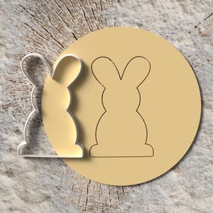 Könnte beinhalten: Ein klarer, hasenförmiger Ausstecher aus Kunststoff neben einem beigefarbenen Kreis mit einer Hasenkontur. Die Artikel befinden sich auf einer hellgrauen und braunen strukturierten Oberfläche, was auf ein Back- oder Bastelthema hindeutet.