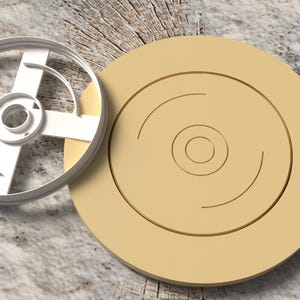 Könnte beinhalten: Ein weißer Metall-Ausstecher mit Kreuzdesign und ein beiger kreisförmiger Keks. Der Keks hat konzentrische Kreise und einen Mittelkreis. Die Artikel befinden sich auf einer Holzmaserungsoberfläche.