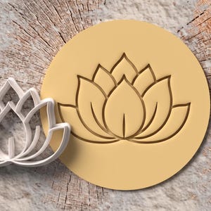 Puede incluir: Un cortador de galletas de plástico blanco con forma de flor de loto junto a un disco circular de masa de galleta de color amarillo claro que presenta un diseño de flor de loto grabado con pétalos simétricos y un detalle de hoja central.