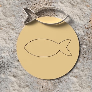 Könnte beinhalten: Ein silberner Ausstecher in Fischform liegt über einem hellgelben Kreis mit einer Fischkontur. Die Objekte befinden sich auf einer strukturierten, hellfarbigen Oberfläche mit einem Holzgrund.