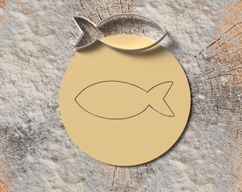 May include: A silver fish-shaped cookie cutter rests above a light yellow circle with a fish outline. The objects are on a textured, light-colored surface, with a wooden background.