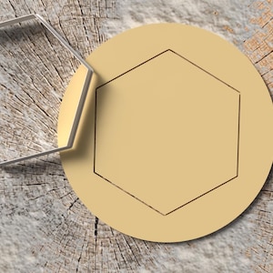 Puede incluir: Un cortador de galletas hexagonal de metal plateado junto a un círculo de color beige con una hendidura hexagonal. Los objetos están sobre una superficie de vetas de madera.