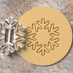 May include: A white plastic snowflake cookie cutter positioned next to a circular piece of light yellow dough that has been embossed with a matching snowflake design, featuring six symmetrical branches with pointed ends.