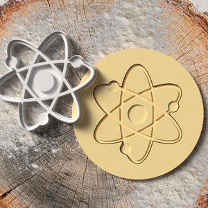 May include: A silver-colored atom-shaped cookie cutter next to a light yellow cookie with the same atom design. The items are on a rustic, wooden surface, suggesting a science or baking theme.