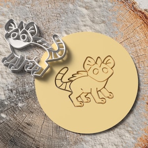 Peut inclure: Un emporte-pièce en métal et un motif estampé d'une créature de dessin animé avec de grandes oreilles et une queue rayée. L'emporte-pièce est argenté et jaune, et le motif estampé est sur un cercle jaune.