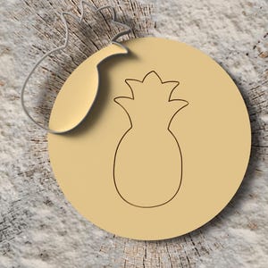 Könnte beinhalten: Hellbeige, kreisförmiger Anhänger mit einer Ananas-Umrisslinie. Der Anhänger hat eine kleine, angebrachte Schlaufe und einen blattförmigen Akzent. Der Hintergrund ist ein strukturiertes, hellbraunes Holz.