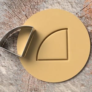 Peut inclure: Un emporte-pièce en métal en forme de quart de cercle à côté d'une découpe de pâte circulaire jaune pâle présentant un motif de quart de cercle en relief. L'emporte-pièce a une finition argentée et la pâte est lisse et plate.