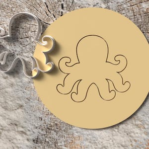 May include: A silver octopus-shaped cookie cutter next to a tan, circular imprint of an octopus. The design is on a wooden surface with a light-colored, textured background.