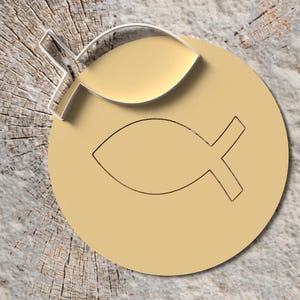 Puede incluir: Un emblema circular dorado con un símbolo de pez, con un cortador de galletas en forma de pez a juego en la parte superior. El emblema y el cortador están sobre un fondo de madera clara texturizada.