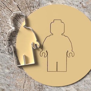 May include: A cookie cutter and a tan circle with a Lego-style figure outline. The cookie cutter is silver and has a shadow. The figure outline is brown. The background is a light brown, textured surface.