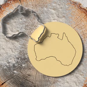 Può includere: Un tagliabiscotti argentato a forma di Australia accanto a un biscotto giallo chiaro con il contorno nero del continente australiano. Gli oggetti sono posti su una superficie di legno.