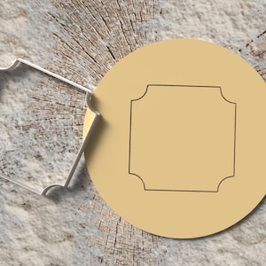 Peut inclure: Un emporte-pièce et un pochoir rond sont présentés sur une surface en bois. L'emporte-pièce est de forme carrée argentée avec des bords décoratifs. Le pochoir est de couleur beige clair avec un contour carré noir.