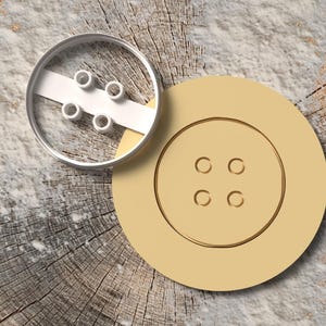 Può includere: Un tagliabiscotti a forma di bottone color beige con quattro fori, accanto a un biscotto con lo stesso design a bottone. Il tagliabiscotti è in metallo e il biscotto è su una superficie di legno.