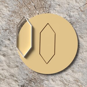 Può includere: Un oggetto circolare beige chiaro con un design geometrico. Un oggetto a forma di diamante argentato, aperto sui lati, è posizionato sopra il cerchio. Una forma a diamante simile è incisa sulla superficie del cerchio. Lo sfondo è un legno marrone chiaro strutturato.